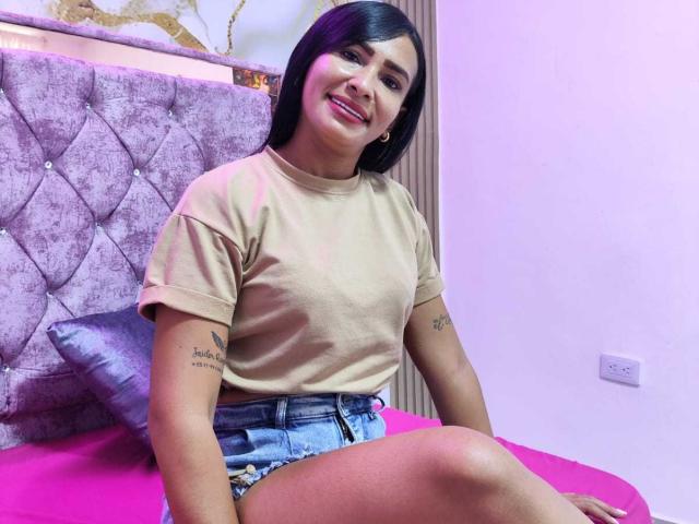 KataleyaHope with black hair, smiling, wearing a beige shirt and denim shorts, showing a tattoo on her right arm, sitting on a pink bed. Boobs are small. Pussy is not visible.