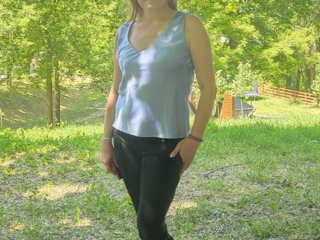 LeaMat has a slim, blue V-neck shirt showing her small tits, black pants, and a wrist bracelet, standing in a grassy, sunlit area.