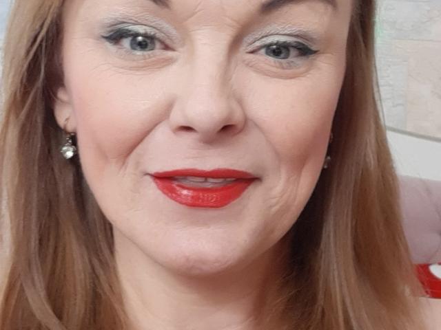 LeaMat's face with red lipstick, light brown hair, blue eyes, and subtle makeup. She's wearing diamond earrings, smiling slightly, with a light skin tone.