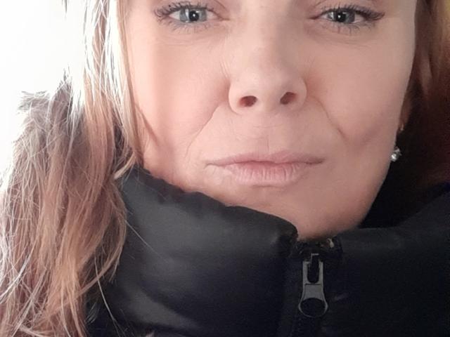 LeaMat's face, showing wrinkles and light brown hair, with blue eyes, black puffy jacket zipper, and subtle smile. Her nose is centered, lips slightly puckered.