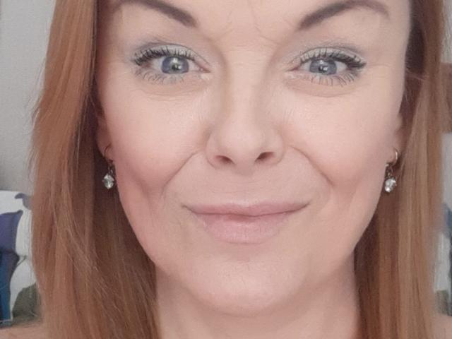 LeaMat has blue eyes, light makeup, and a slight smile with her red hair down, wearing small diamond earrings, and showing a subtle, natural-looking pussy.