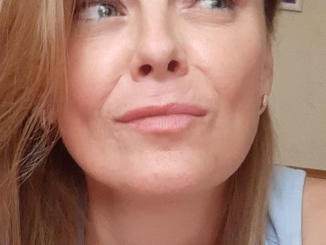 LeaMat's face, showing a slight smile with pinkish lips, light brown hair, and blue eyes, wearing a light blue top, with subtle makeup and small earrings.