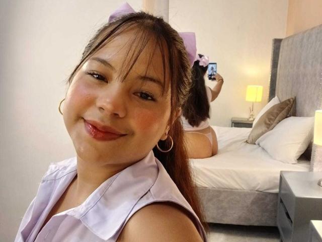 KathiaSmith with puffy pink bow, hoop earrings, smiling, pink shirt, brown hair, pink lips, holding phone, back to camera, on bed, white pillows, gray headboard, yellow lamp.