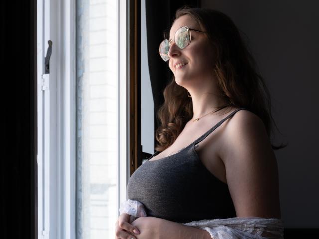 CoralieLaCoquine with glasses, smiling at sunlight, wearing a gray tank top, showing her tits, and a white lace shirt off her shoulders.