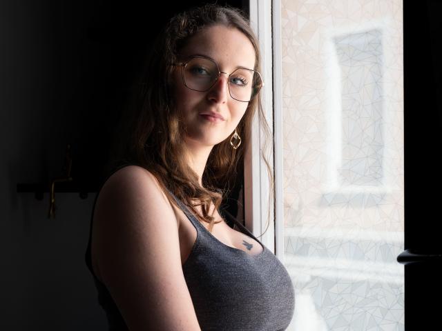 CoralieLaCoquine with glasses, brown wavy hair, gray tank top, showing her tits, standing by a window, subtle smile, light coming from outside, wearing gold hoop earrings.