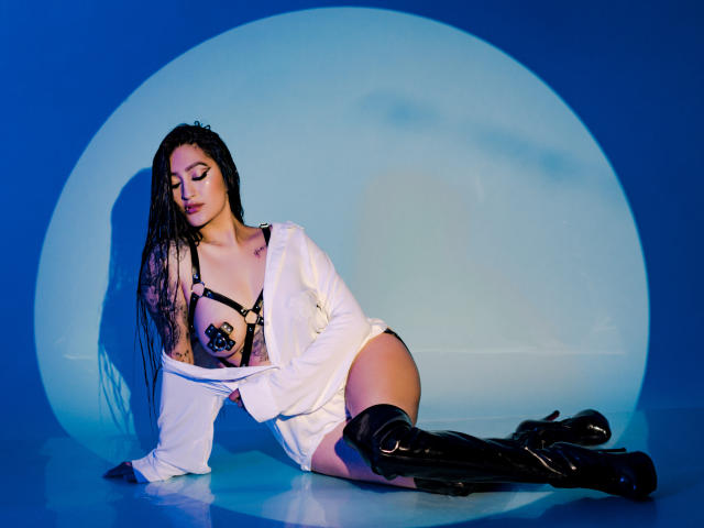 EmmaRosseti with long black braids, wearing an open white shirt, black harness with star pasties, and black knee-high boots, kneeling on blue-lit floor, showing her big ass.