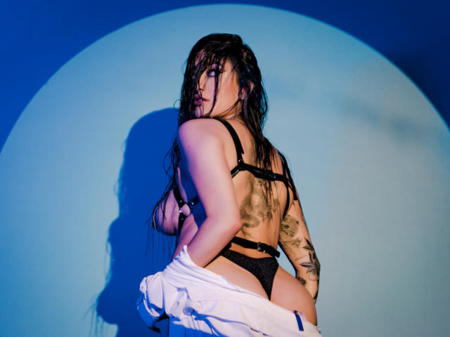 EmmaRosseti with wet black hair, wearing black bra and panties, white jacket pulled down, showing her ass and tattoos, looking back, shadow on blue wall, sexy.