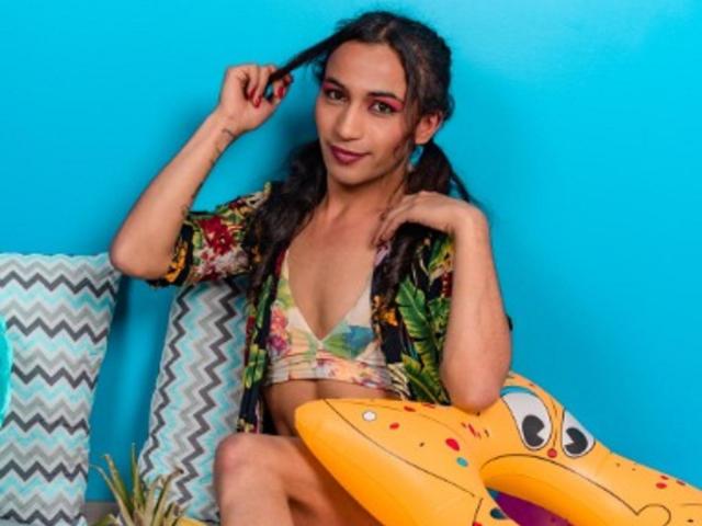 GisselyTs with pigtails, wearing a floral bra and open shirt, holding hair, smiling, and sitting on a yellow inflatable with a cute face, against a bright blue wall.
