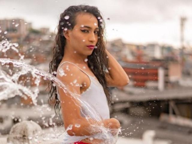 GisselyTs with wet, curly black hair, wearing a white, wet, see-through tank top, standing in a water splash, looking to the side with pink lipstick.