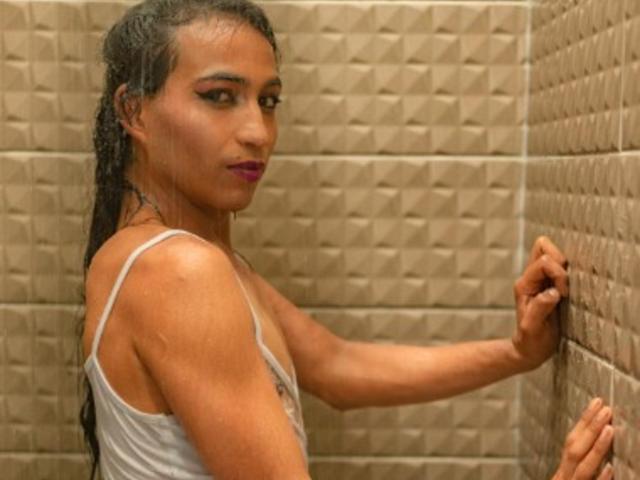 GisselyTs with wet hair, wearing a white tank top, standing in a tiled shower, looking over her shoulder, touching the wall with one hand, showing her ass.