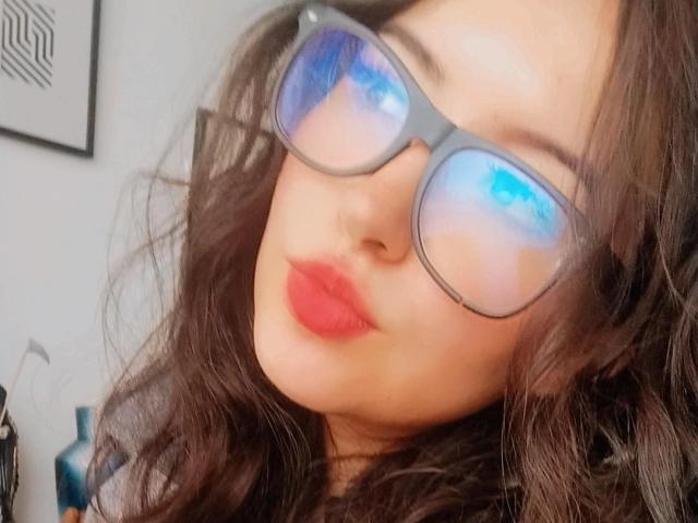 SusanMonroy with blue-tinted glasses, red lips, and wavy dark brown hair, looking slightly to the side. Her nose is small and her skin is light.