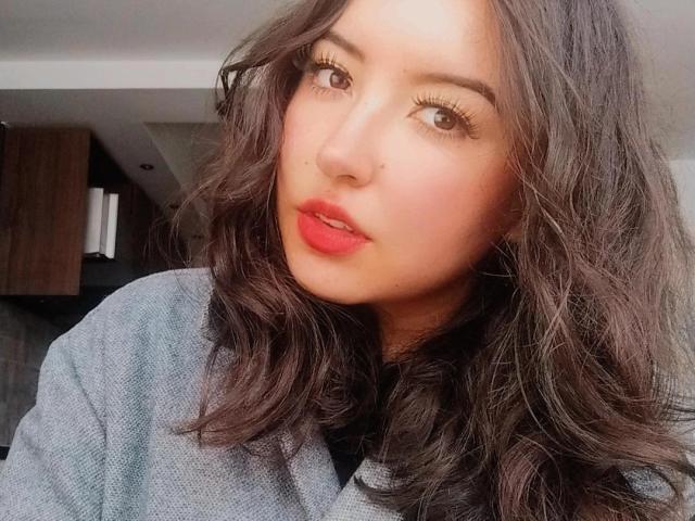 SusanMonroy has wavy brown hair, red lipstick, and long eyelashes. She's wearing a gray sweater and looking at the camera with a slight pout. Her tits are not visible.