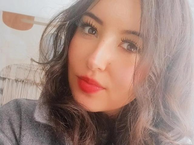 SusanMonroy's face, with wavy dark brown hair, red lipstick, and subtle makeup, wearing a gray textured shirt. She has large brown eyes and a slight, confident smile.