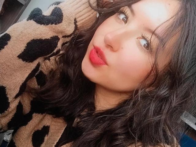 SusanMonroy with wavy black hair, wearing a beige and black leopard print sweater, has red lipstick, long eyelashes, and a cute, slightly pouty expression.