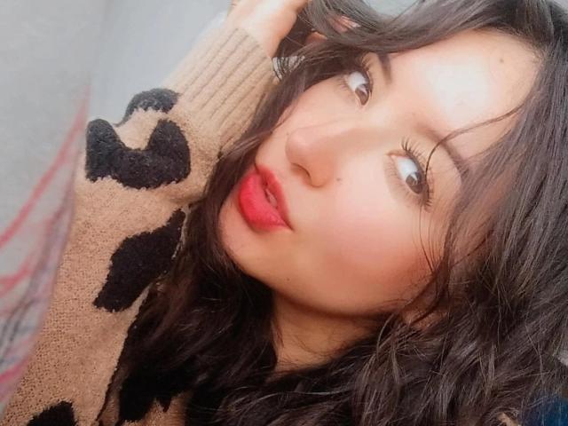 SusanMonroy with wavy black hair, red lipstick, and light brown eyes, wearing a beige sweater with black patterns, looking up with a slight smile. Her hand is in her hair.