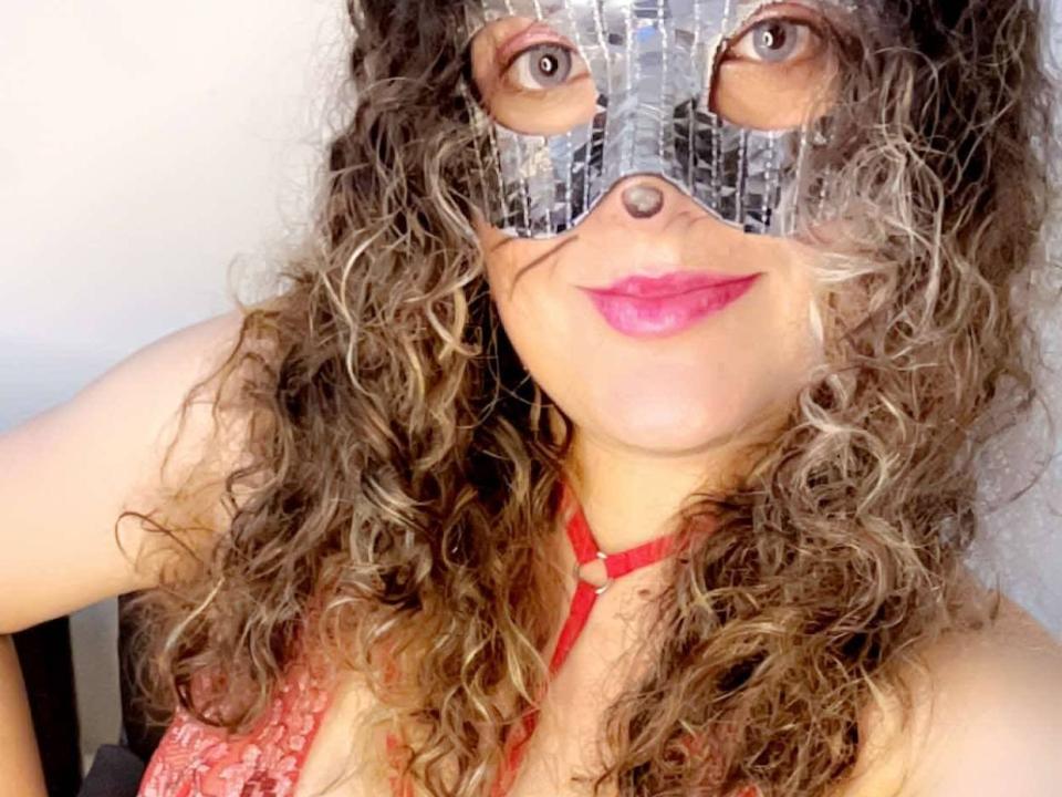 Curly-haired LuanaWinner with pink lips, wearing a metallic mask covering her eyes, and a red halter top, smiling at the camera. Her tits are partially visible.
