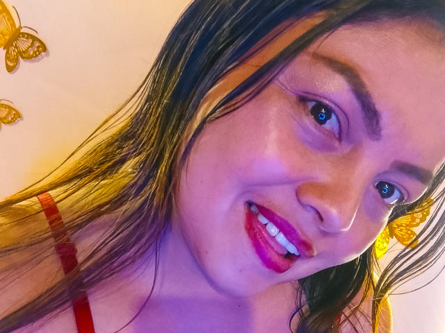 Kharo with wet brown hair, red lipstick, and purple eyeshadow, smiling at the camera, wearing a red top, with yellow butterflies on the left.