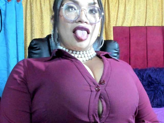 MarieStanFord with glasses, tongue out, wearing a pearl necklace and a maroon cardigan that shows her large tits, sitting in a black leather chair.