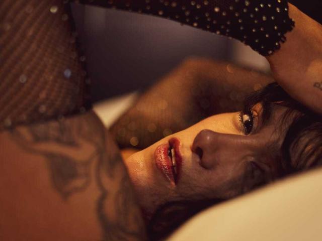 ElektraKizz lying on a bed with dark fishnet stockings and a tattooed arm, gazing up with parted red lips, wearing a black, sparkly top, in dim, warm light.