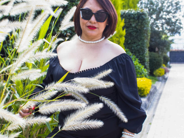 KaroMichelle with dark red bob, black off-shoulder dress, pearl necklace, red lipstick, and black sunglasses, holding fluffy grass, showing her tits. Outdoor setting, daytime.