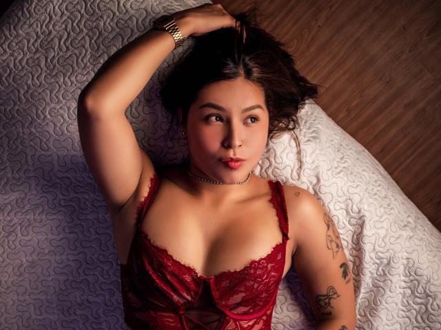 Abby Thompson ligt op een bed, gekleed in rode kant lingerie, met haar hand in haar bruine haar, waarbij tatoeages op haar arm zichtbaar zijn, en draagt een choker ketting, en kijkt naar de camera..
