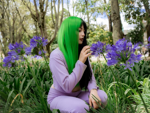 AngelBlakee with green hair, purple outfit, crouching, smelling purple flowers in a garden. She's got black hair, cute, and her tits are slightly visible under the long-sleeve top.
