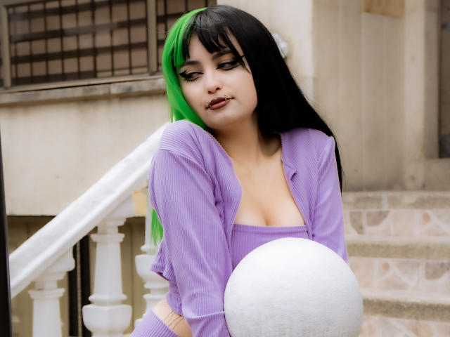 AngelBlakee, met groen en zwart haar, draagt een paars gestreept topje en een vest, en houdt een witte bal vast. Ze heeft volle lippen en een enigszins verveelde uitdrukking..