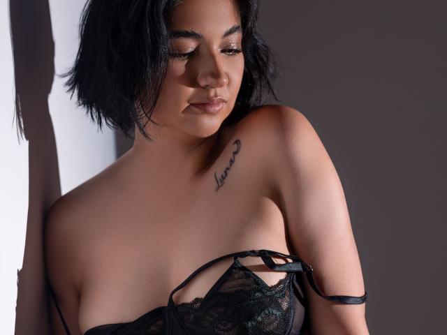 MiaWallaceOh with short black hair, wearing black lace lingerie, showing her tits and a tattoo of "Love" on her left shoulder, eyes closed, and a soft, confident expression.