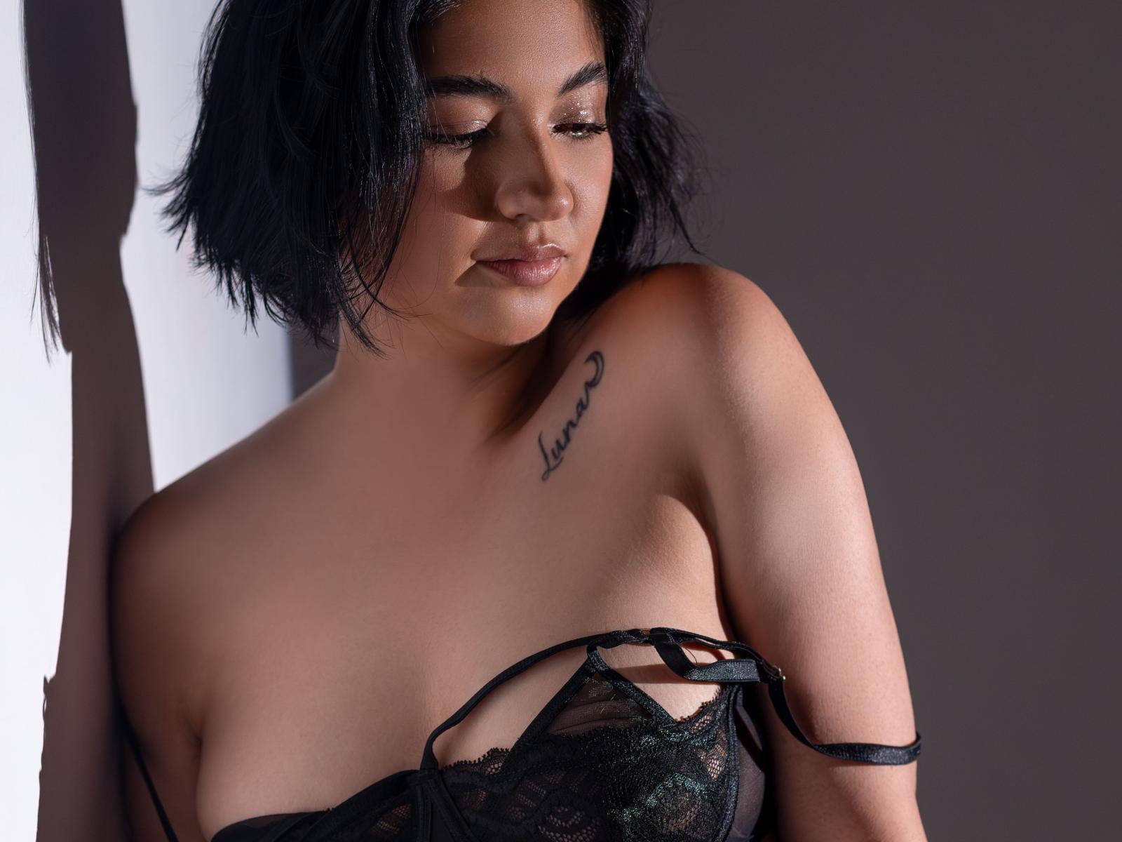 MiaWallaceOh with short black hair, wearing black lace lingerie, showing her tits and a tattoo of "Love" on her left shoulder, eyes closed, and a soft, confident expression.