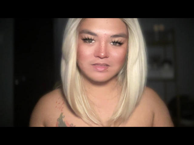 GoddessAnitaX with blonde hair, green eyes, and a nose piercing, showing off her small tits and a colorful tattoo on her right shoulder. Blurred dark background.