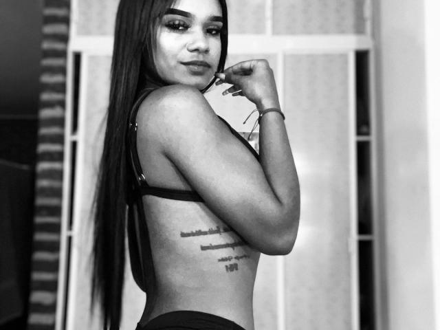 Black-and-white photo of IsabellaLov with long straight hair, looking over shoulder, wearing a bra and holding a phone, showing a tattoo on her side. Her tits are visible.
