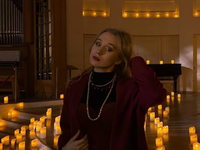 AstraMluse, avec ses cheveux blonds, portant un blazer rouge, un pull noir en col roulé et un collier de perles, se tient parmi des bougies allumées, la main dans les cheveux, regardant légèrement sur le côté, dans une pièce faiblement éclairée..