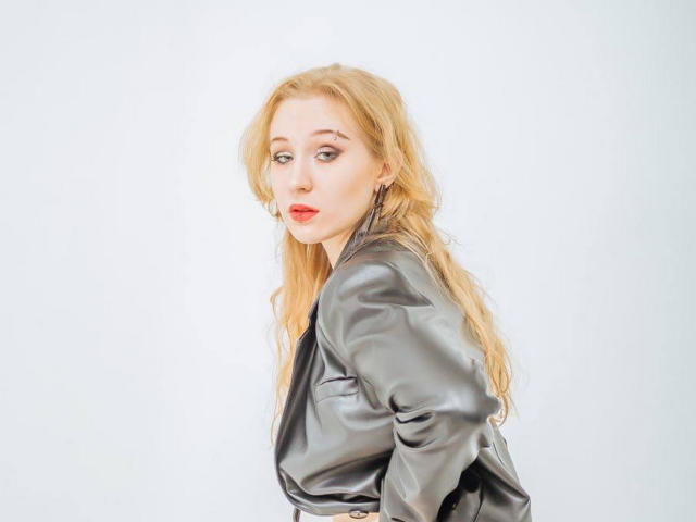 Eine Frau mit langen, blonden Haaren, die eine glänzende schwarze Lederjacke trägt, blickt zurück, mit rotem Lippenstift, heller Haut und leichtem Make-up, die Arme hinter dem Rücken..