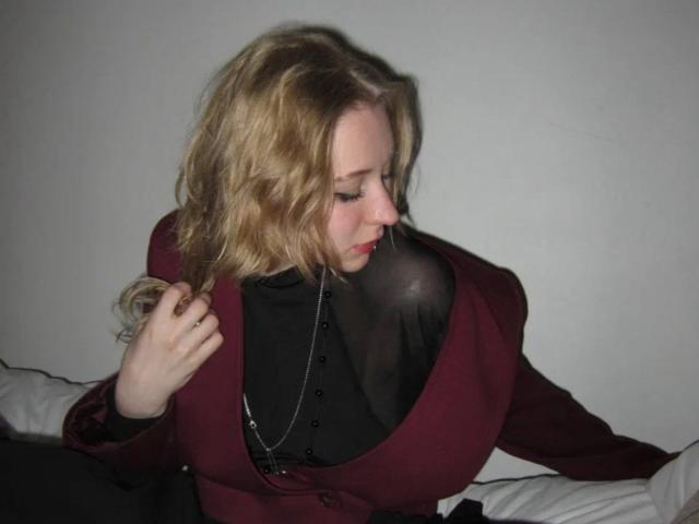 Une femme nommée AstraMluse, avec des cheveux blonds ondulés, portant un haut marron et une chemise noire transparente, laissant entrevoir de larges seins, regardant vers le bas, une main dans ses cheveux, sur un lit blanc..