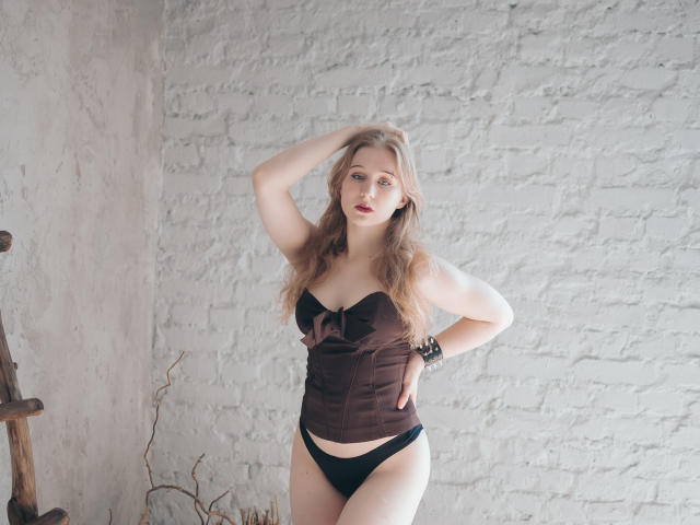 Eine Frau mit langen, blonden Haaren, die ein braunes Korsett und schwarze Unterwäsche trägt, mit einer Hand auf dem Kopf, die in die Kamera schaut, vor einer weißen Ziegelwand, stehend, weiches Licht, dezentes Make-up, Armband am Handgelenk..