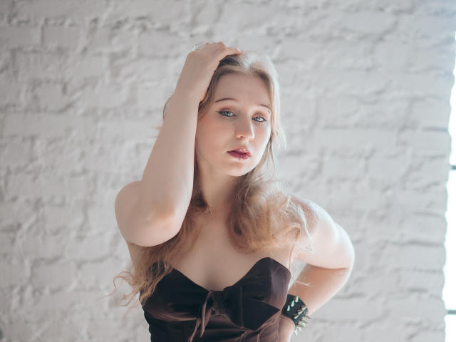 Astra, avec ses cheveux blonds, sa peau claire et ses yeux bleus, portant une robe noire sans bretelles, une main dans les cheveux, regardant légèrement sur le côté, avec une expression subtile et séduisante..