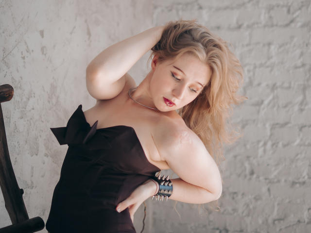 Eine Frau mit blonden Haaren, die ein schwarzes, trägerloses Kleid trägt, berührt ihren Kopf, hat die Augen geschlossen, rote Lippen, einen silbernen Armreif, eine weiße Mauer aus Ziegelsteinen im Hintergrund, weiches Licht, einen entspannten Gesichtsausdruck und helle Haut..