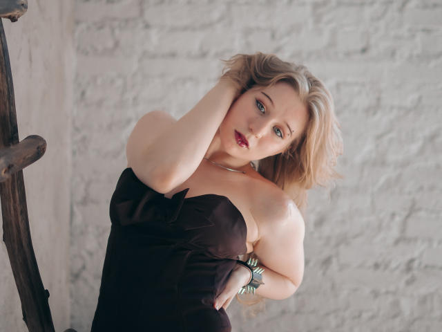 Astra, une femme aux cheveux blonds, portant une robe noire sans bretelles, une main dans les cheveux, les yeux bleus, du rouge à lèvres, un bracelet au poignet droit, debout près d'une échelle en bois..