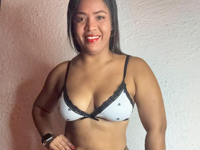 AliceBooty, con seni marroni medi, indossa un reggiseno bianco con pizzo nero, sorride con rossetto rosso, porta un orologio nero ed è in piedi di fronte a un muro con una superficie strutturata..