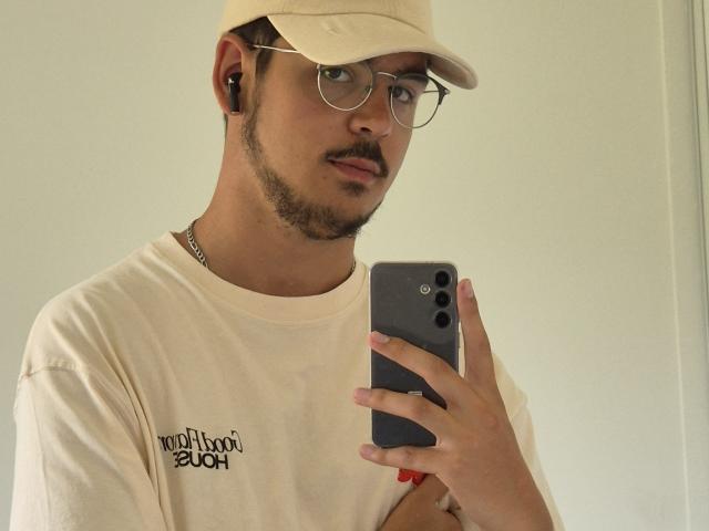 LeonardStone with a beige cap, glasses, and mustache, wearing a cream shirt, holding a gray phone, looking into the camera, earbuds in.