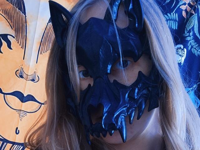 NellyFreda with a black, glossy wolf mask covering her face, blonde hair, and blue-tinted skin, looking up with serious eyes. She's wearing the mask in a selfie.