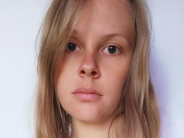 NellyFreda's face with pale skin, blonde hair, green eyes, and pinkish lips, looking slightly to the side with a neutral expression. Her nose is small and slightly upturned.