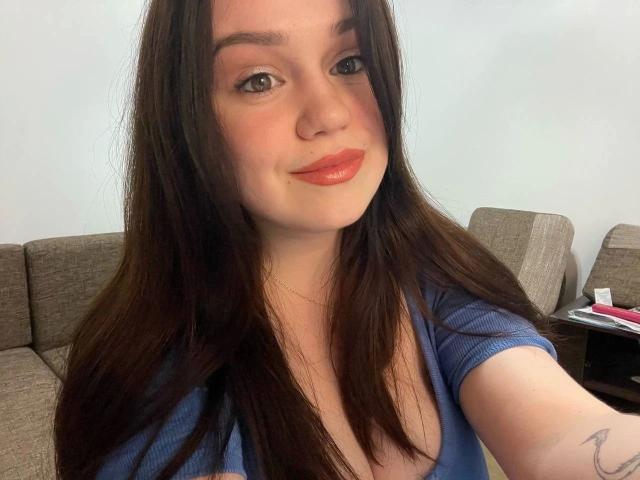 SweetVibe-hot with long brown hair, wearing a blue top, showing a cute smile with orange lipstick, and a small tattoo on her right arm. Boobs slightly visible in the shot.