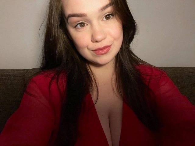 SweetVibe-hot with long brown hair, wearing a red V-neck shirt, showing her cleavage, smiling softly, brown eyes, light skin, sitting on a gray couch.