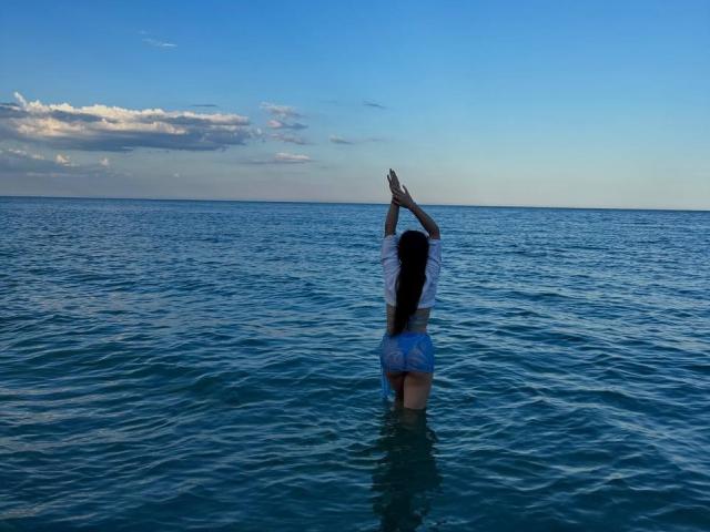 PaigePrado with long dark hair, arms raised, wearing a white top and blue shorts, standing waist-deep in clear blue water, facing away.