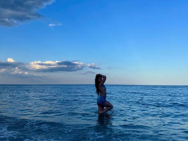 PaigePrado, standing in shallow blue water, wearing a purple bikini, her ass slightly raised, long brown hair, arms behind head, clear sky, small clouds.