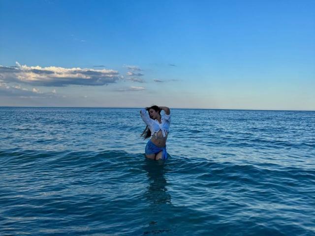 PaigePrado with long dark hair, white long-sleeve shirt, and blue shorts, standing waist-deep in clear blue water, arms raised, hands in hair, under a bright blue sky.