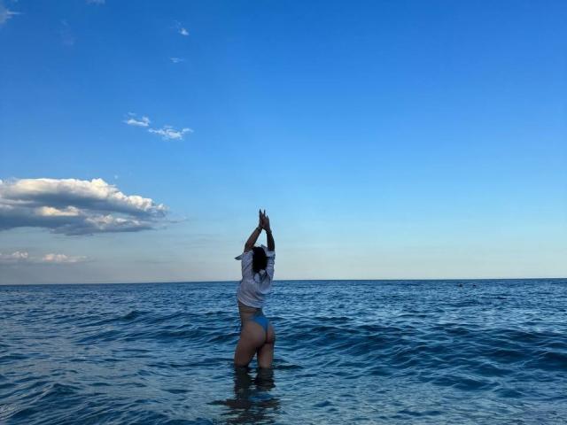PaigePrado stands in blue ocean water, arms raised, wearing a white shirt and blue thong, showing her ass and back to the camera, under a clear blue sky.