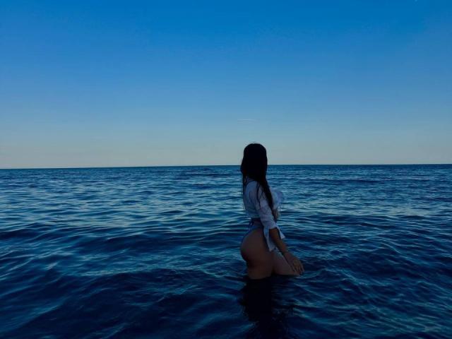PaigePrado's ass is in the water, wearing a white shirt, with her back to the camera, in a blue ocean under a clear sky. Her pussy is not visible.
