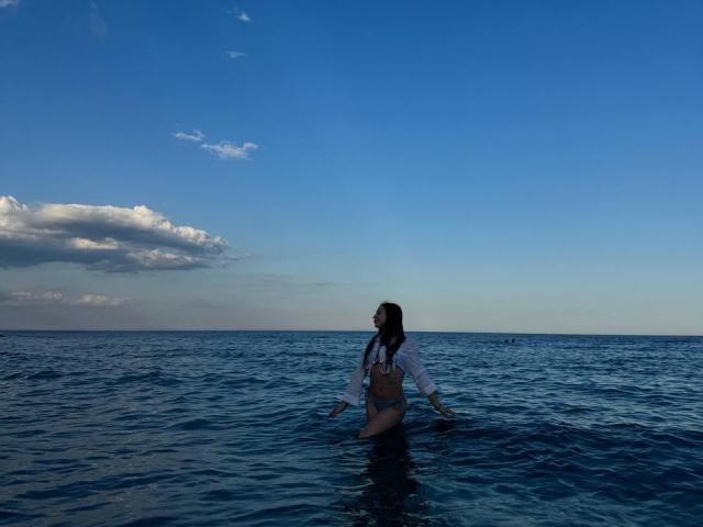 PaigePrado in a blue ocean, wearing a white shirt and brown bikini bottom, with long dark hair, standing in water, arms relaxed, under a clear blue sky.